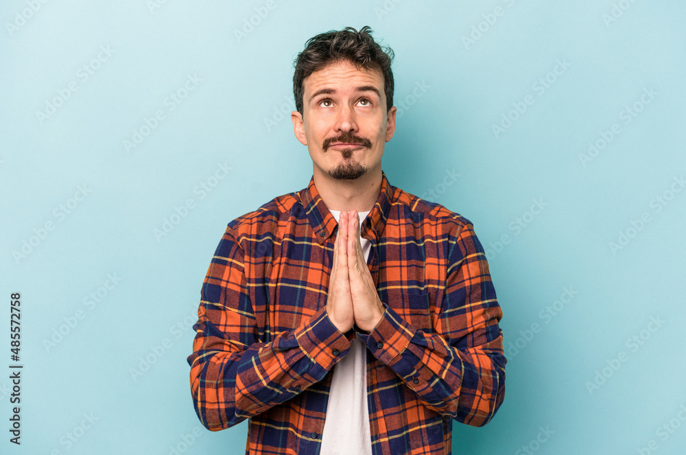 Young caucasian man isolated on blue background holding hands in pray near mouth, feels confident. Young caucasian man isolated on blue background holding hands in pray near mouth, feels confident.