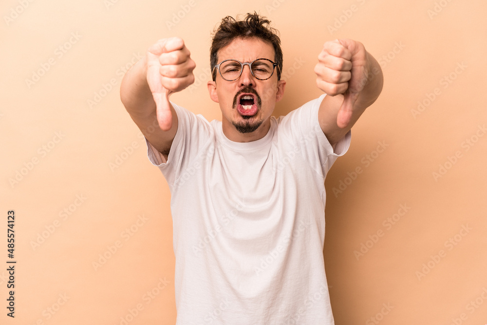 Young caucasian man isolated on beige background showing thumb down and expressing dislike. Young caucasian man isolated on beige background showing thumb down and expressing dislike.