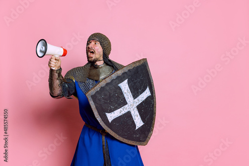 Portrait of man, medieval warrior or knight in armor with shield shouting in megaphone isolated over pink studio background