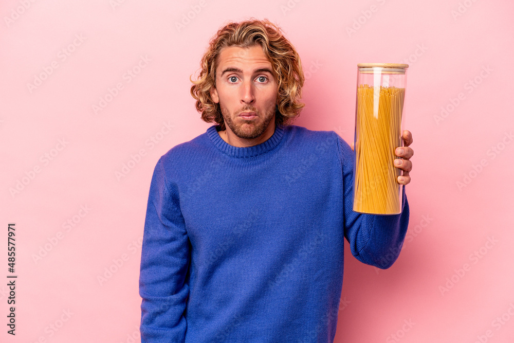 Young caucasian man holding spaghettis jar isolated on pink background shrugs shoulders and open eyes confused. Young caucasian man holding spaghettis jar isolated on pink background shrugs shoulders and open eyes confused.