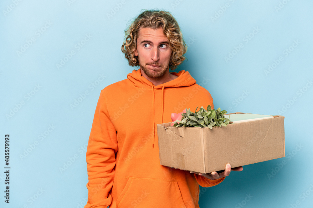 Young caucasian man making a move while picking up a box full of things isolated on blue background confused, feels doubtful and unsure. Young caucasian man making a move while picking up a box full of things isolated on blue background confused, feels doubtful and unsure.