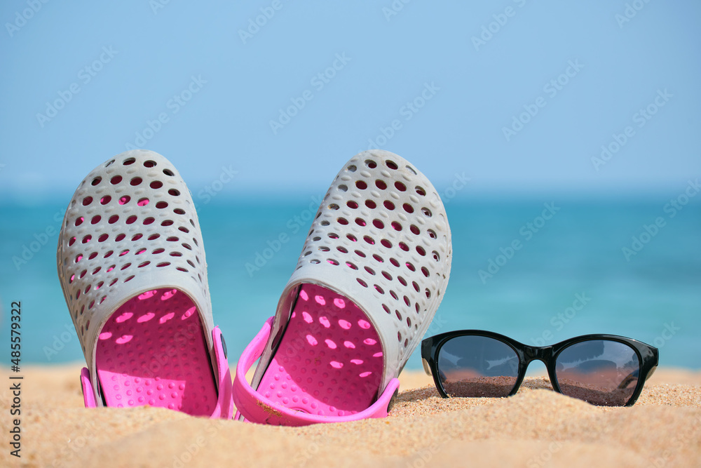 Obraz premium Closeup of clogs shoes and black protective sunglasses on sandy beach at tropical seaside on warm sunny day. Summer vacation concept