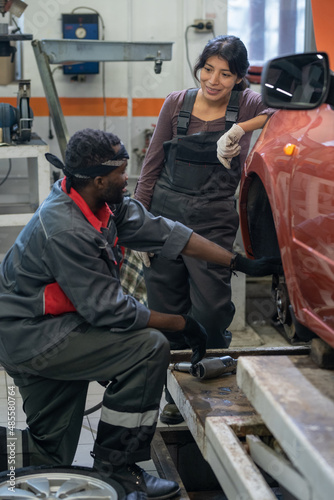 Vertical portrait of two ethnic mechanics repairing car in garage shop, focus on smiling young woman wearing workwear