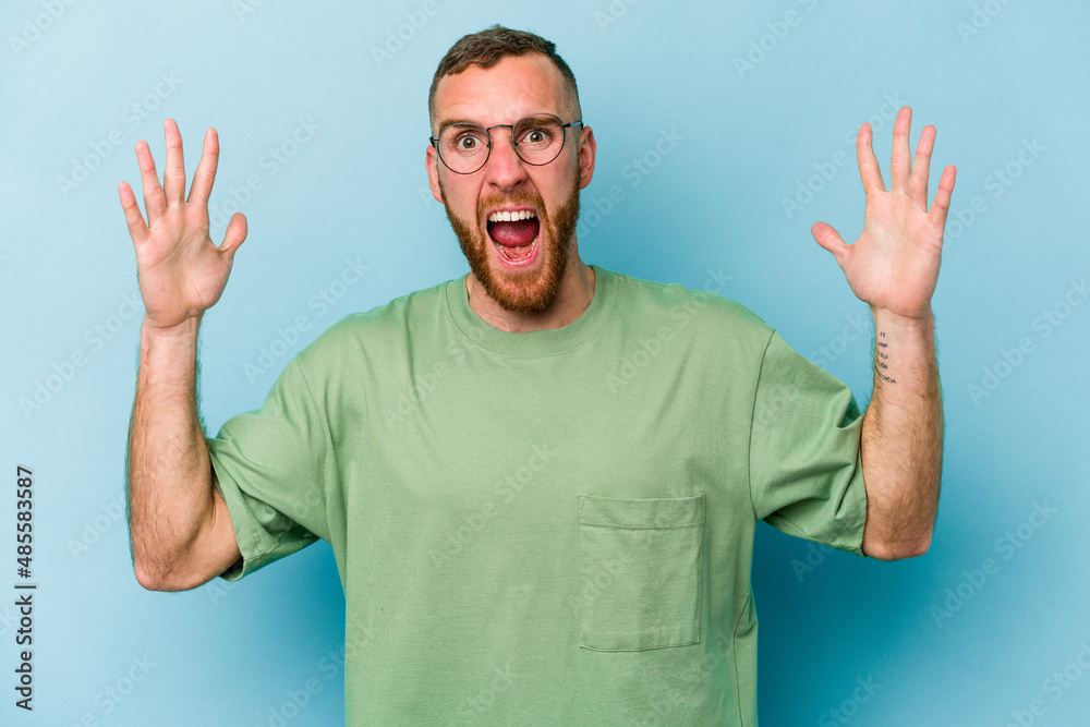 Naklejka premium Young caucasian man isolated on blue background receiving a pleasant surprise, excited and raising hands.