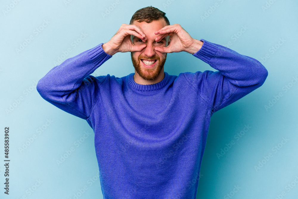 Young caucasian man isolated on blue background showing okay sign over eyes Young caucasian man isolated on blue background showing okay sign over eyes