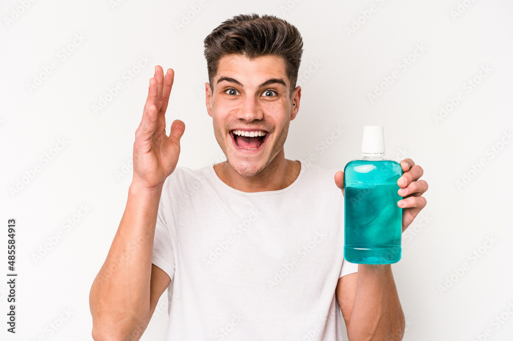Young caucasian man holding mouthwash isolated on white background receiving a pleasant surprise, excited and raising hands. Young caucasian man holding mouthwash isolated on white background receiving a pleasant surprise, excited and raising hands.