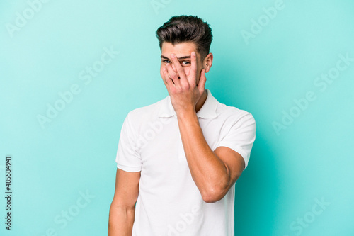 Young caucasian man isolated on blue background blink at the camera through fingers, embarrassed covering face.