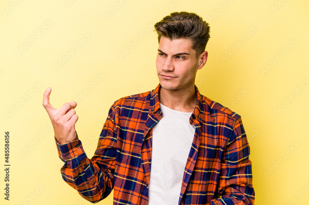 Young caucasian man isolated on yellow background pointing with finger at you as if inviting come closer. Young caucasian man isolated on yellow background pointing with finger at you as if inviting come closer.