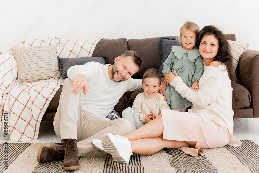 Stylish happy parents sitting near the sofa with joyful little kids, smiling, spending weekends at home. Charming father with pretty mother playing with cute daughter and son, family concept Stylish happy parents sitting near the sofa with joyful little kids, smiling, spending weekends at home. Charming father with pretty mother playing with cute daughter and son, family concept