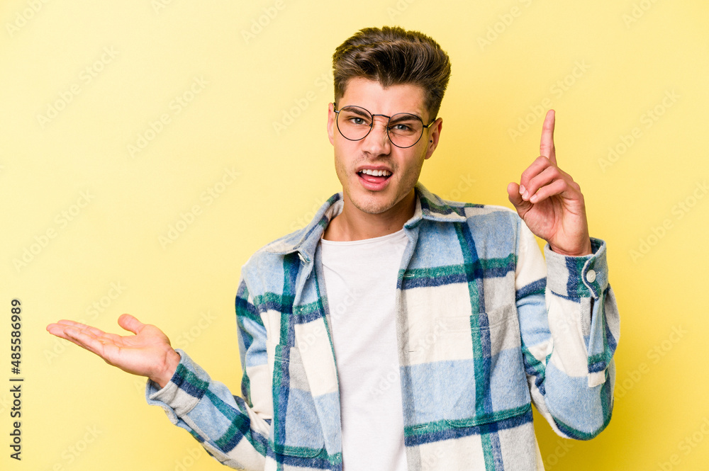 Young caucasian man isolated on yellow background holding and showing a product on hand. Young caucasian man isolated on yellow background holding and showing a product on hand.