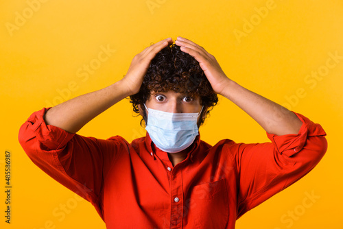 Oh my God. Portrait of shocked young man in casual style with surgical medical mask standing, holding head and looking at camera with unbelievable face. Indoor studio shot, isolated on orange