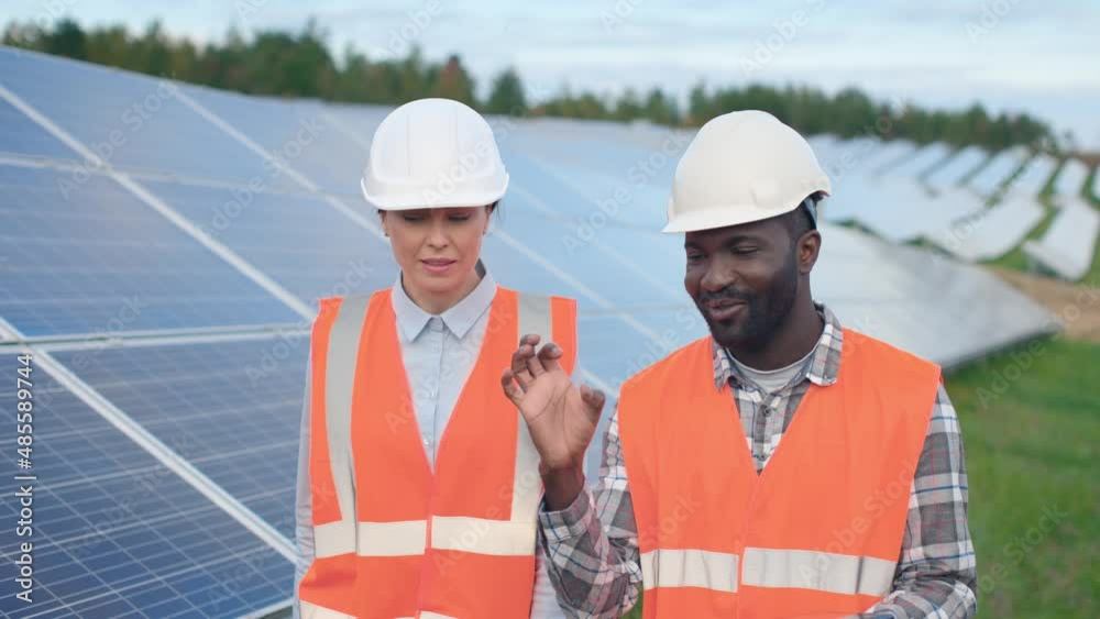 Close-up experienced African-American worker walks among field of solar panels with Caucasian female colleague and discusses specifics of professional tasks. Close-up experienced African-American worker walks among field of solar panels with Caucasian female colleague and discusses specifics of professional tasks.