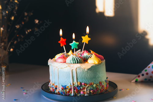 Colorful birthday cake with sprinkles and burning star shaped candles on a dark wall background with shadows. Festive birthday celebration, party. Selective focus, copy space