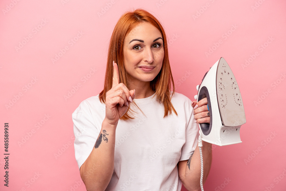 Young caucasian woman holding iron isolated on pink background showing number one with finger. Young caucasian woman holding iron isolated on pink background showing number one with finger.