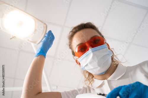 Close-up of a young woman dentist in red ultraviolet protective glasses, wears a medical mask, the dentist treats her teeth. POV of the patient.