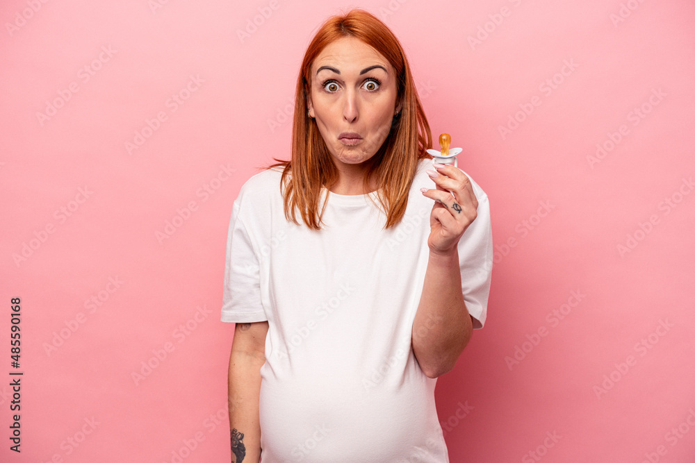 Young caucasian pregnant woman holding pacifier isolated on pink background shrugs shoulders and open eyes confused. Young caucasian pregnant woman holding pacifier isolated on pink background shrugs shoulders and open eyes confused.