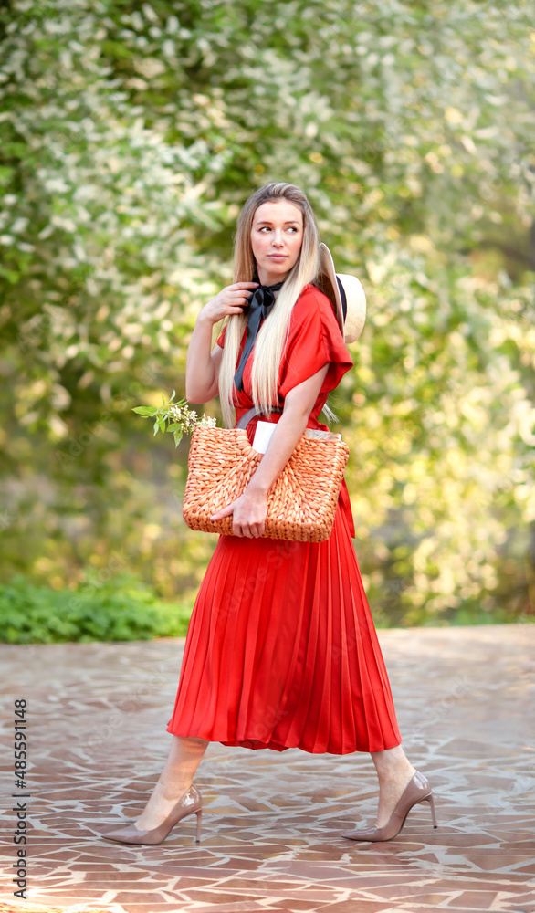 Businesslike young woman in a bright red dress, hat and high-heeled shoes walks through a blooming park. In her hands she has a wicker summer bag with documents. Businesslike young woman in a bright red dress, hat and high-heeled shoes walks through a blooming park. In her hands she has a wicker summer bag with documents.