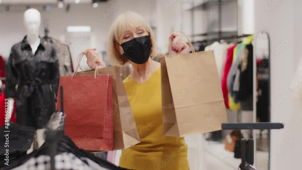Happy middle-aged woman in protective mask standing in clothing store holding bags in hands dynamically moves dances rejoices in purchases bought at discount on sale new collection satisfied customer Happy middle-aged woman in protective mask standing in clothing store holding bags in hands dynamically moves dances rejoices in purchases bought at discount on sale new collection satisfied customer