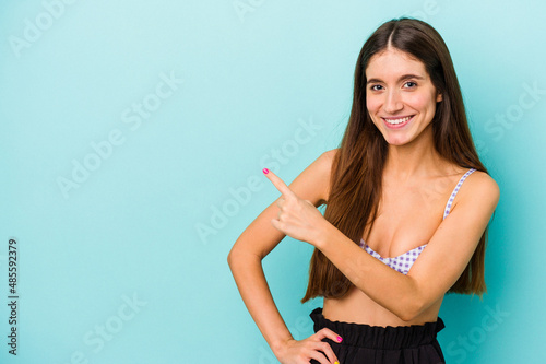 Young caucasian woman wearing bikini isolated on blue background smiling and pointing aside, showing something at blank space.