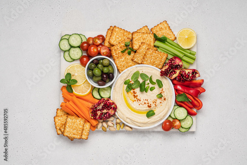 Hummus plate with vegetable snacks. Carrot, cucumber, tomato, celery sticks, dry crackers, olives and hummus bowl on marble tray and light background, top view