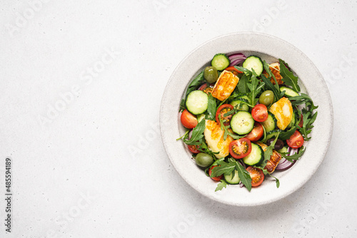 Halloumi salad with arugula, tomato, cucumber, onion and olives in bowl on light background. Mediterranean dish with fried cheese and vegetables