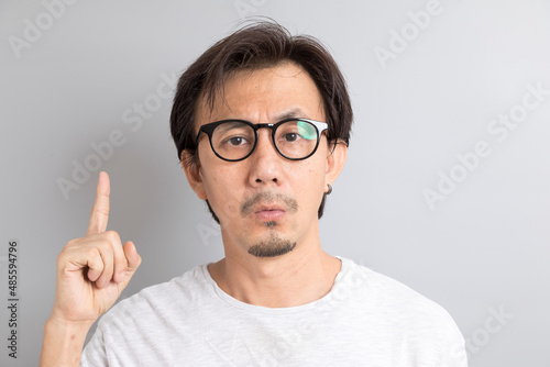 Adult asian man with glasses pointing fingers upward with curious face on grey background.
