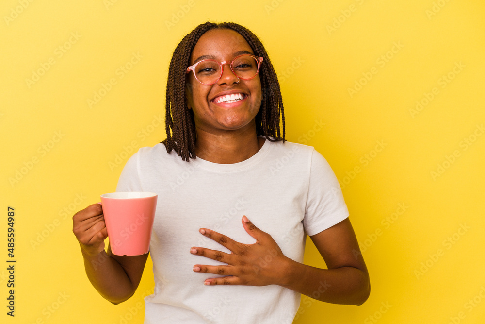 Young african american woman holding a mug isolated on yellow background laughs out loudly keeping hand on chest. Young african american woman holding a mug isolated on yellow background laughs out loudly keeping hand on chest.