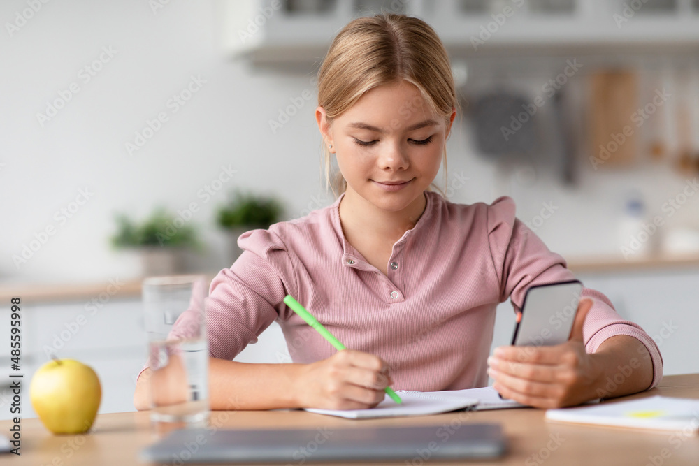 Serious pretty blonde european teenager girl makes notes with smartphone at table in minimalist kitchen Serious pretty blonde european teenager girl makes notes with smartphone at table in minimalist kitchen