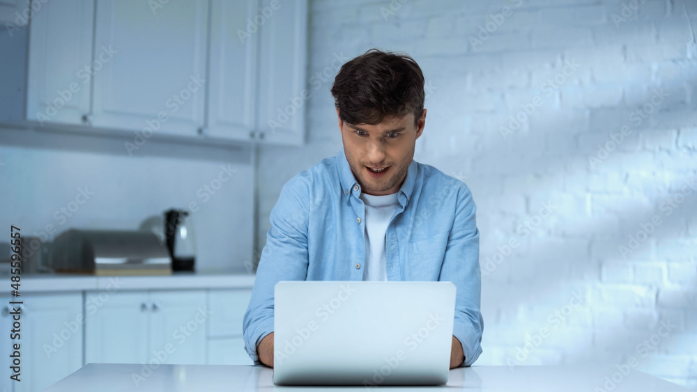 astonished freelancer in blue shirt working on laptop in kitchen astonished freelancer in blue shirt working on laptop in kitchen