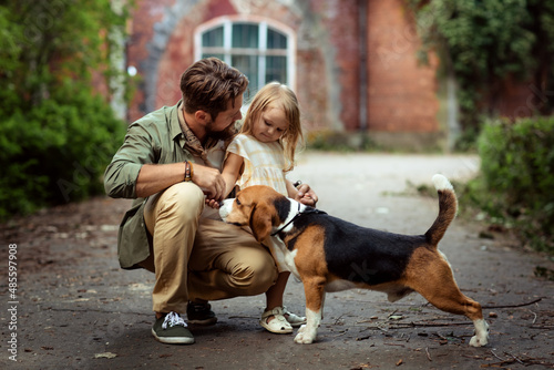 Caucasian father playing with small daughter and pet dog beagle in casual clothes  in front of medieval brick mansion in park. Summer, leisure activity, family conception. Weekend recreation