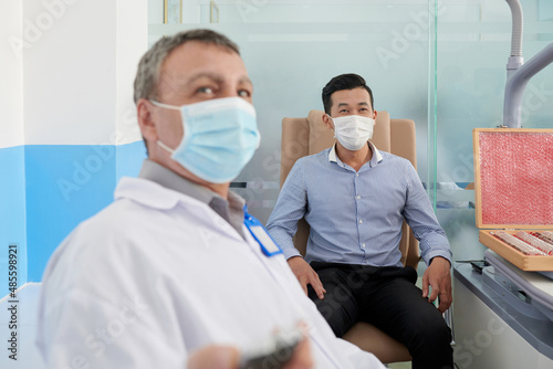 Doctor asking patient in medical mask to read letters on test chart hanging on wall when checking his eyesight