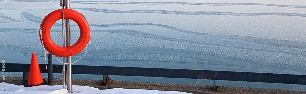 An orange lifesaver on the harbour with water surface texture of melting ice and ripples.  Winter banner background