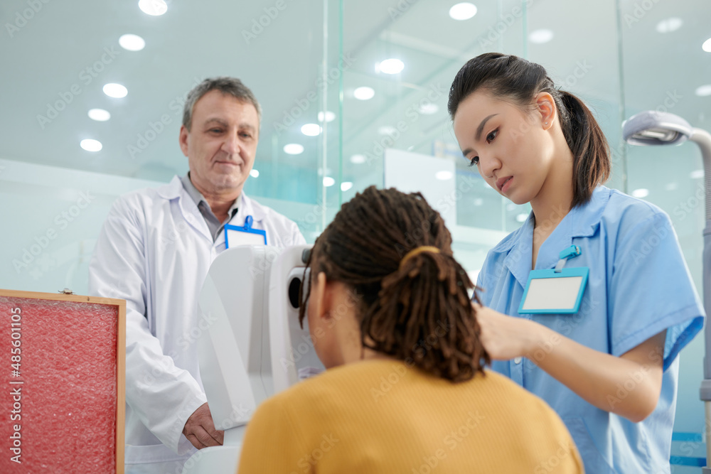 Nurse helping patient to put head in autorefractor so the doctor could measure the refractive error of the eyes and prescribe lenses Nurse helping patient to put head in autorefractor so the doctor could measure the refractive error of the eyes and prescribe lenses