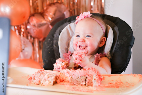 Cute birthday girl celebrating her one year birthday by messily eating a cake surrounded by pink background streamers and balloons