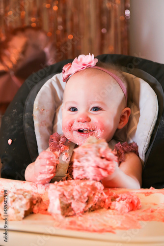 Cute birthday girl celebrating her one year birthday by messily eating a cake surrounded by pink background streamers and balloons