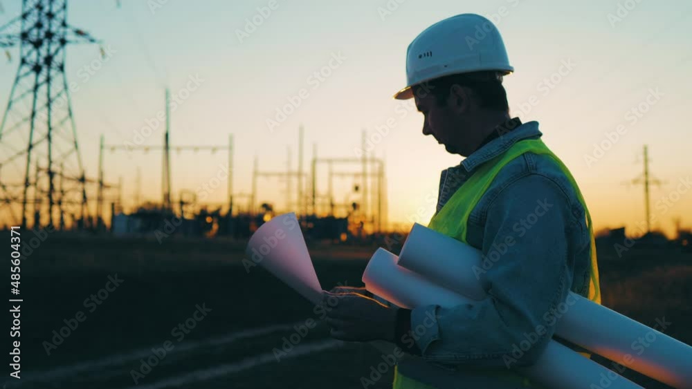 Power work concept. Engineer standing on field with electricity towers. Young engineer putting security helmet. Electrical engineer with high voltage electricity pylon at sunset background.