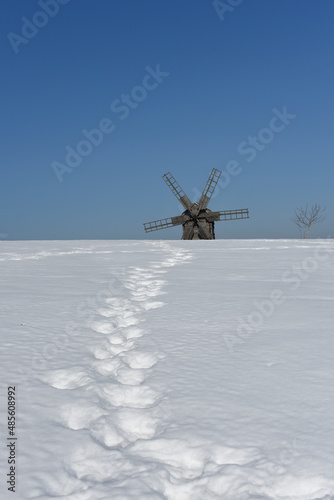 A winter landscape with a mill on the horizon and deep human footprints leading to it in a snowy field. Winter trekking