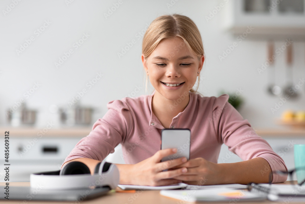 Happy caucasian teenage girl blonde typing on smartphone at table in cozy kitchen interior Happy caucasian teenage girl blonde typing on smartphone at table in cozy kitchen interior