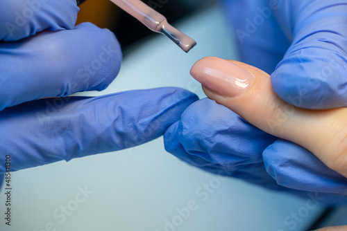 Close-up of a manicure master in blue gloves applies a layer of pink gel polish to the client's nail. Coating the nail plate with nail polish. Professional manicure in the salon close-up.