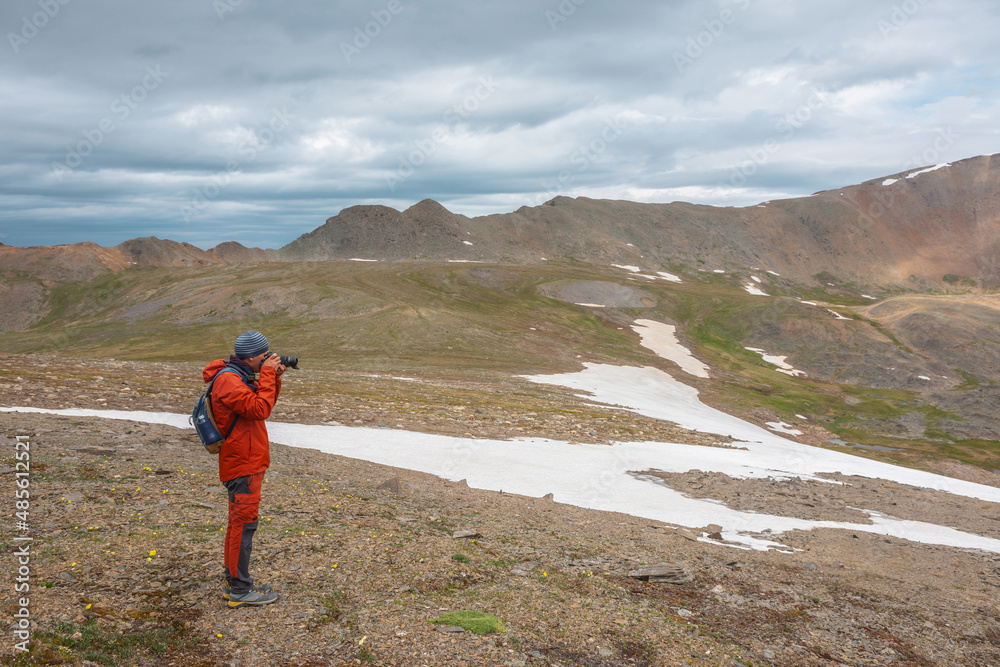 Obraz premium Atmospheric scenery with man in red against mountain range with sharp rocks under gray cloudy sky. Tourist with camera shoots dramatic mountain landscape. Man photographs mountains at gloomy weather.
