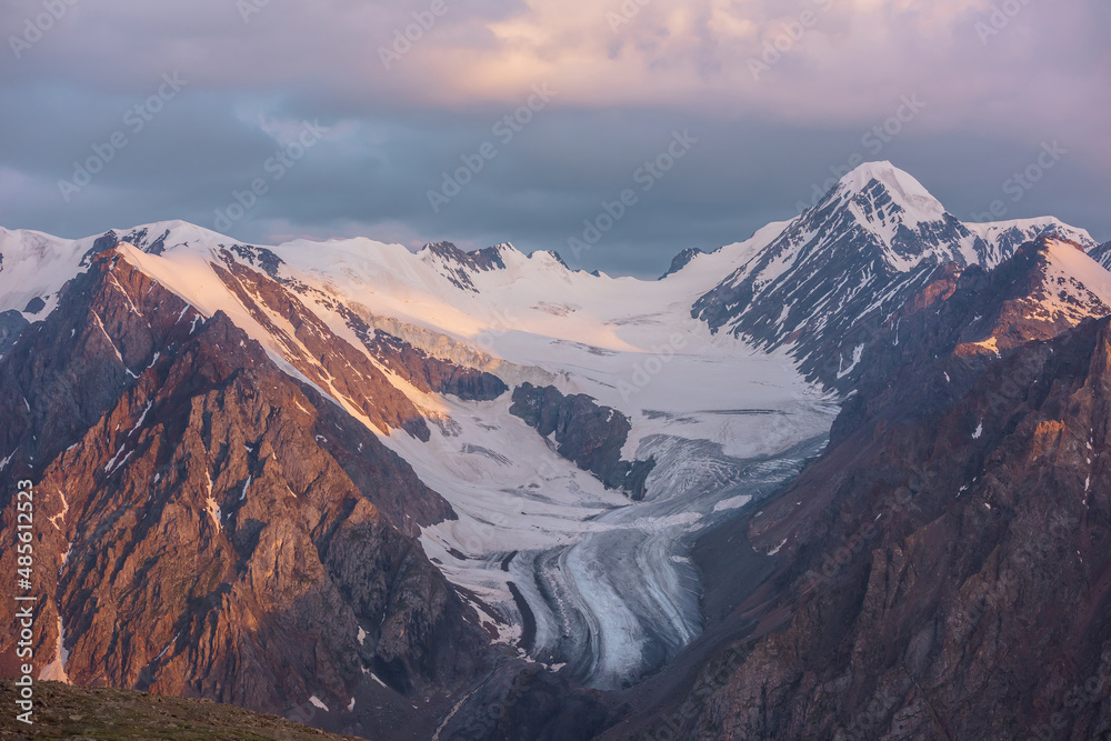 Dramatic aerial view to high snow mountain peak in early morning at dawn. Awesome scenery with sunlit snow mountains in cloudy sky at sunrise. Scenic landscape with large glacier in sunrise colors.