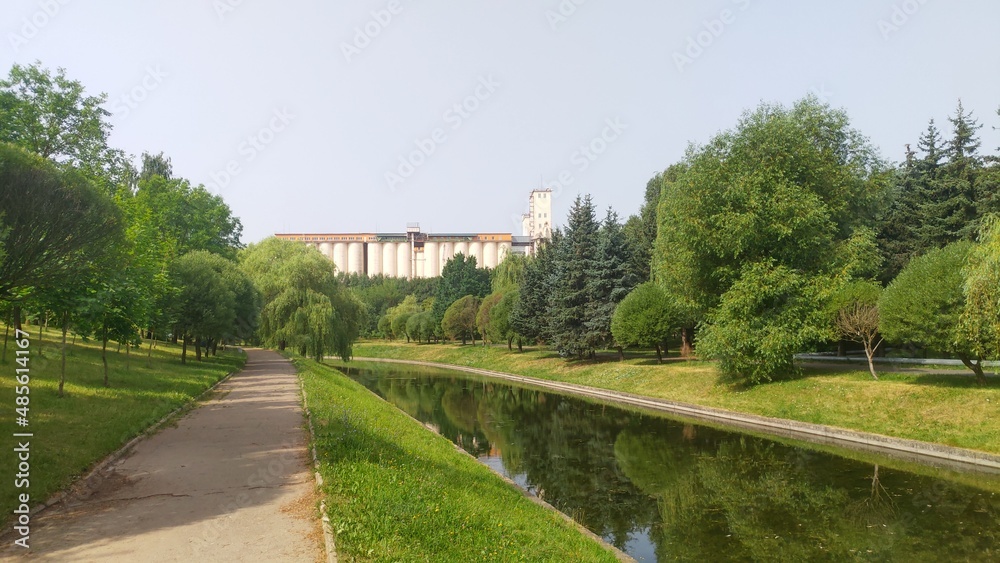 Through the city park among the grassy lawns and trees runs a canal with water. Its banks are concreted. Behind the trees you can see a large grain elevator located outside the park. Sunny weather Through the city park among the grassy lawns and trees runs a canal with water. Its banks are concreted. Behind the trees you can see a large grain elevator located outside the park. Sunny weather