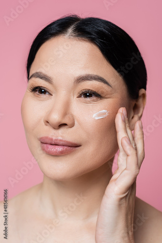 Asian woman applying cream on cheek isolated on pink