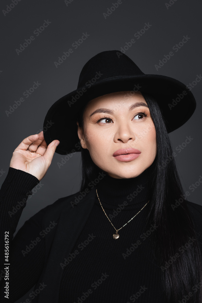 Portrait of stylish asian woman adjusting fedora hat isolated on dark grey Portrait of stylish asian woman adjusting fedora hat isolated on dark grey
