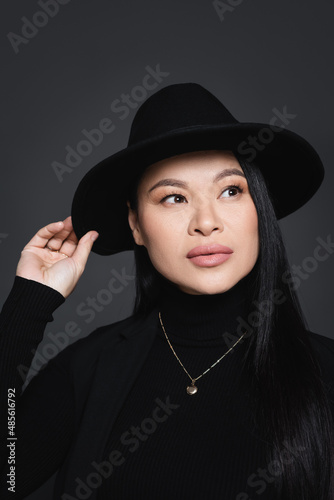 Portrait of stylish asian woman adjusting fedora hat isolated on dark grey © LIGHTFIELD STUDIOS