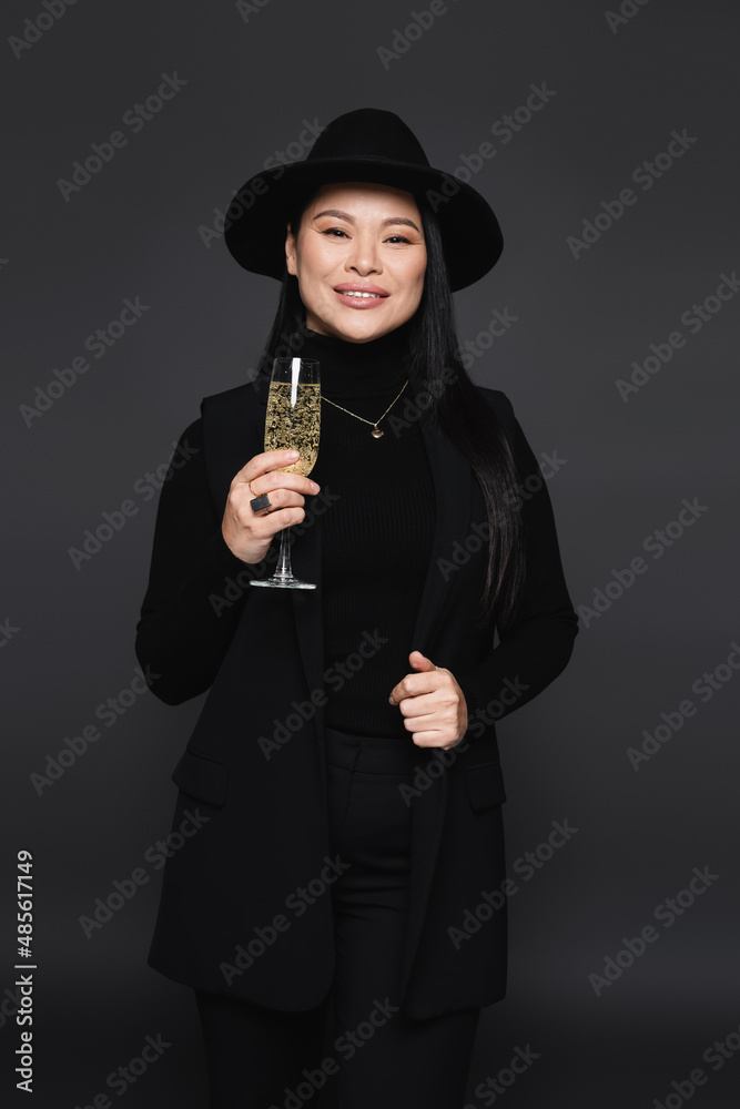 Positive asian woman in fedora hat holding champagne isolated on dark grey Positive asian woman in fedora hat holding champagne isolated on dark grey