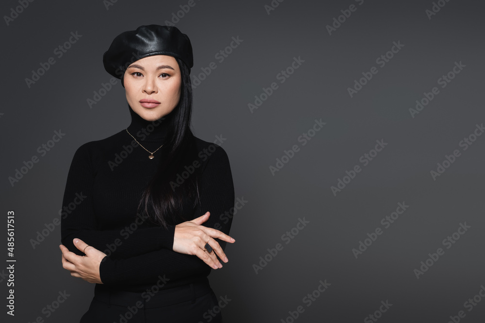 Trendy asian model in beret and necklace looking at camera isolated on dark grey Trendy asian model in beret and necklace looking at camera isolated on dark grey