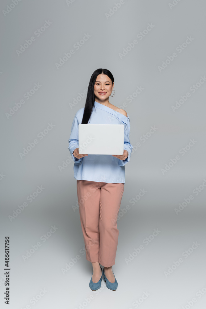 Positive and stylish asian woman holding laptop on grey background Positive and stylish asian woman holding laptop on grey background