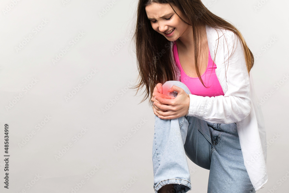 Blonde young girl in teenage wearing having knee pain raised it up and holding with her hands, looks with painful facial expression.