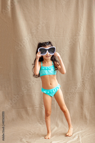 Funny little girl about 6 years old in a casual summer swimsuit posing on a plain beige background. The concept of a child's lifestyle. Layout of the copy space.